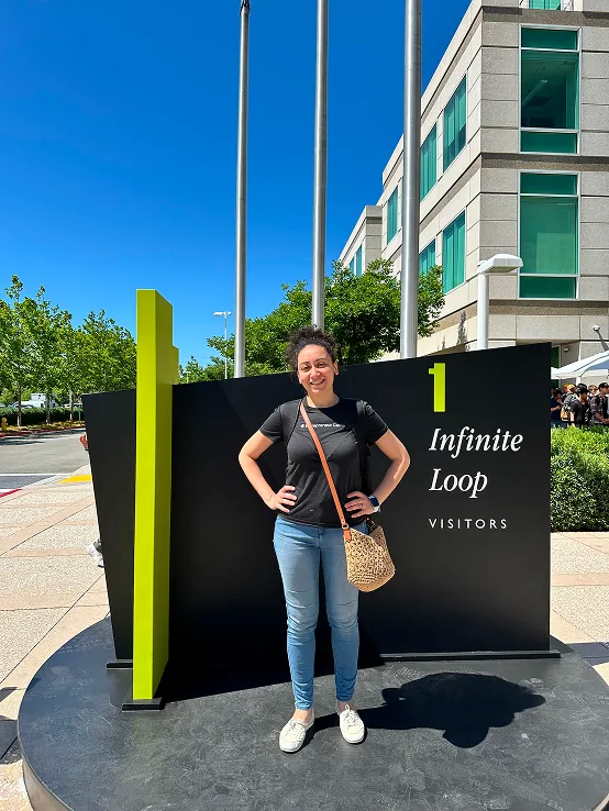 Sylvia Said steht lächelnd neben dem Schild "1 Infinite Loop Visitors" auf Apples Campus in Cupertino