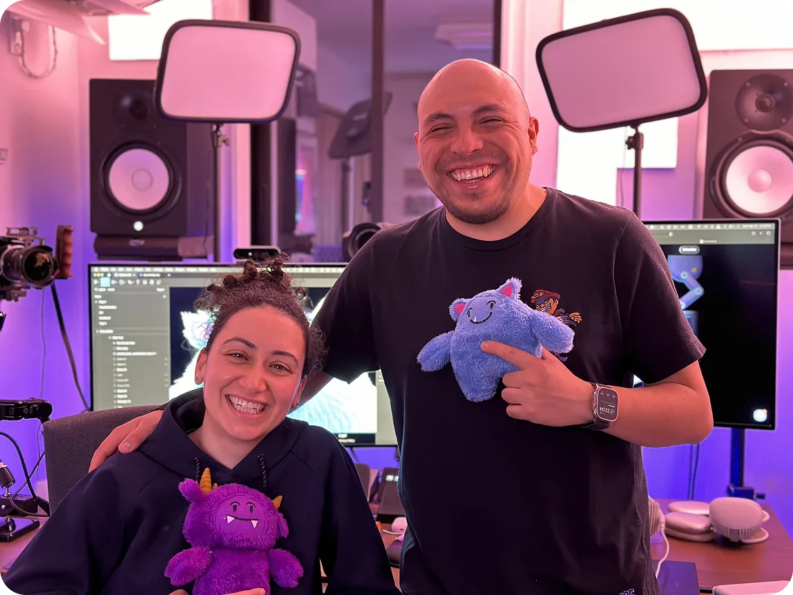Sylvia and Daniel smiling in a studio, holding the purple and blue Arabic4Kids plush characters